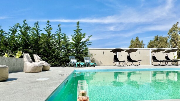 Área da piscina com espreguiçadeiras, uma mesa com uma taça de vinho e uvas, e um céu azul límpido.