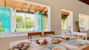 Uma área de refeições com uma mesa posta para refeição, janelas grandes com cortinas e vista para o verde lá fora.