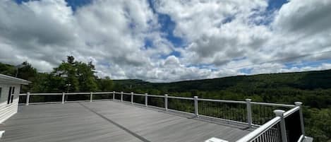 A wooden deck with white railings, overlooking a forested landscape under a partly cloudy sky.