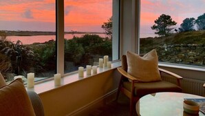 Une pièce dotée d’une grande fenêtre donnant sur un plan d’eau et sur un coucher de soleil. Plusieurs bougies sont posées sur le rebord de la fenêtre, et une chaise avec un coussin s’y trouve.