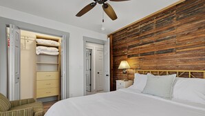 Une chambre d’hôtel comprenant un lit, une table de chevet, une lampe, une tête de lit en bois, une armoire avec des vêtements suspendus et un ventilateur de plafond.