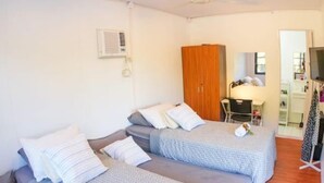 Ein Hotelzimmer mit drei Betten, jeweils mit gemusterten Bettdecken und Kissen. Es gibt einen Holzschrank, einen Schreibtisch mit Stuhl sowie einen an der Wand befestigten Fernseher.