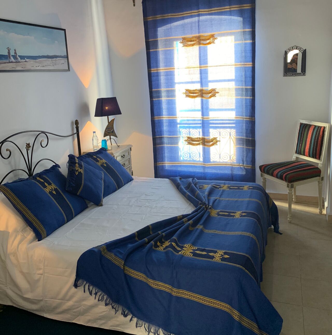 Une chambre à coucher comprenant un lit, une chaise, une table de chevet, une lampe, un tableau et une fenêtre avec des rideaux.