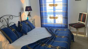 Une chambre à coucher comprenant un lit, une chaise, une table de chevet, une lampe, un tableau et une fenêtre avec des rideaux.