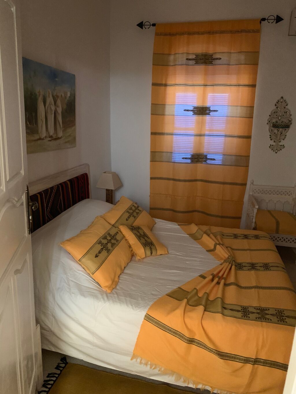 Ein Schlafzimmer mit einem Bett, einem Nachttisch, einer Lampe, einem Bild an der Wand und einem Fenster mit Vorhängen.