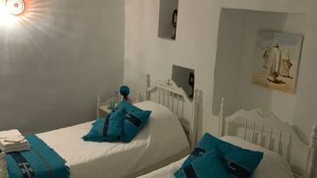A bedroom with two single beds, each with a blue and white duvet, white headboards, and a small bedside table with a lamp.