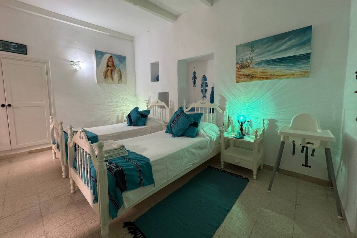 Un dormitorio con dos camas, un armario blanco, una mesita con una lámpara y un cuadro de una playa.