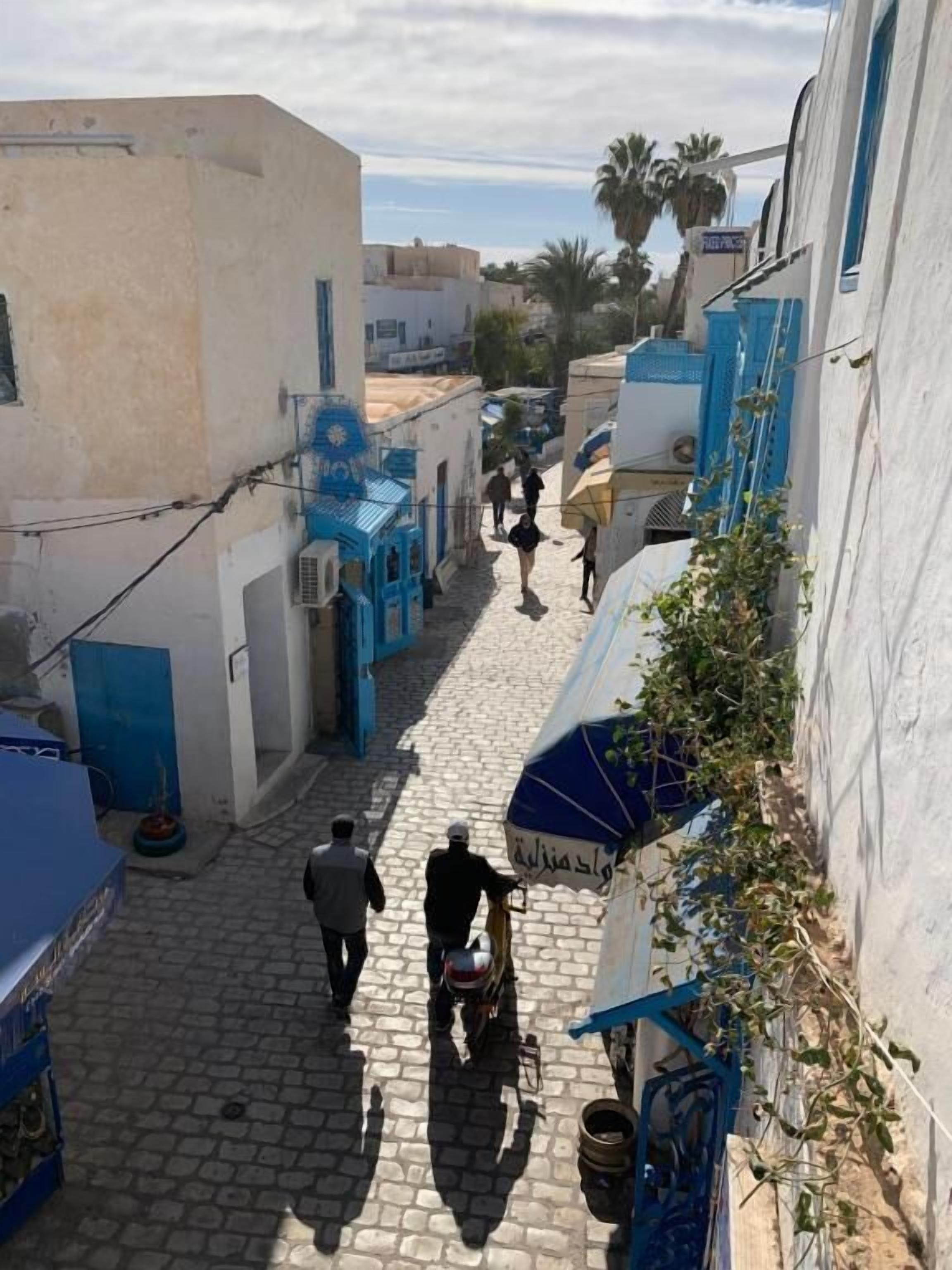 A narrow cobblestone street flanked by buildings with blue accents and a clear sky.