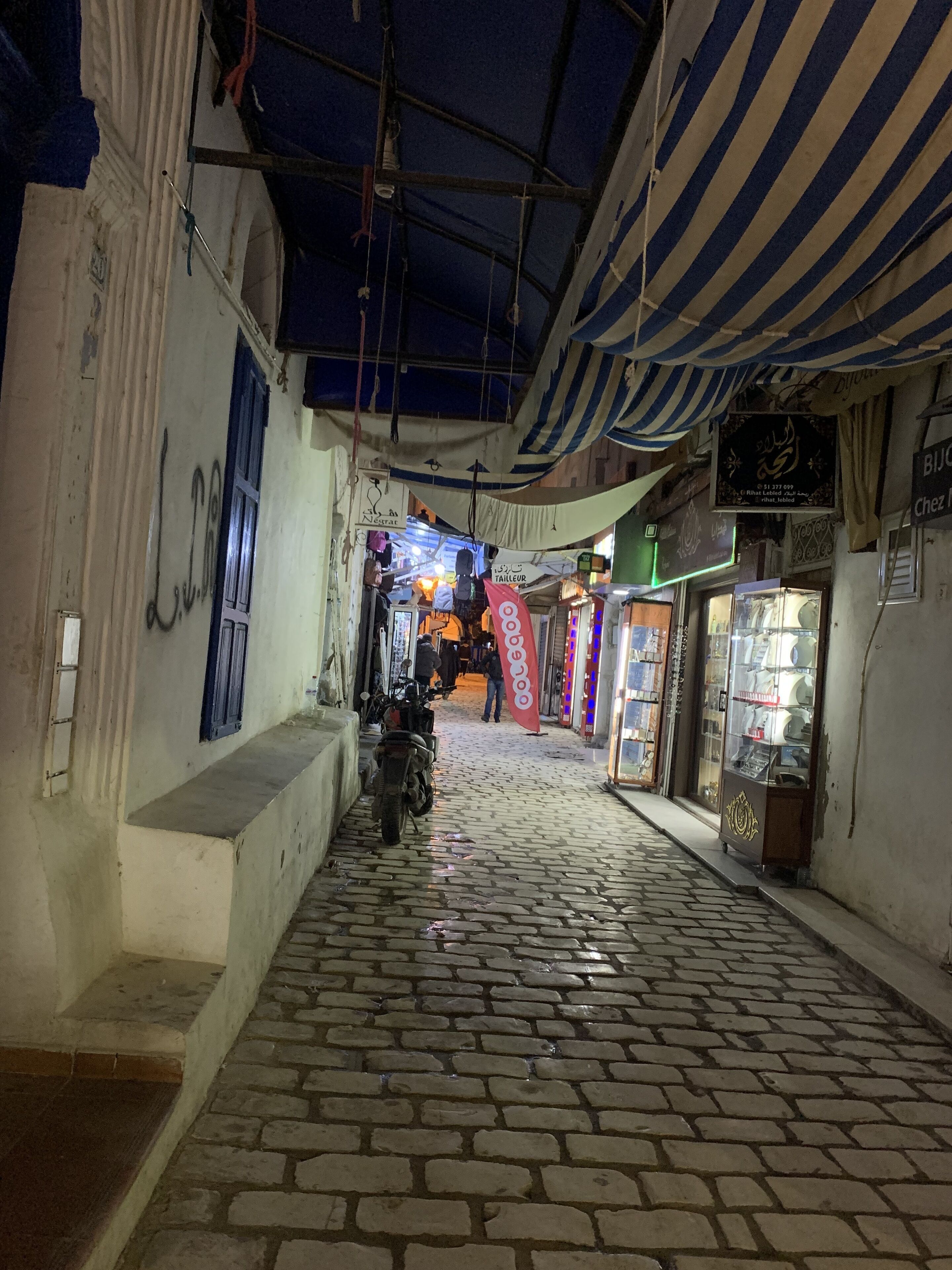 A narrow cobblestone alley with shops on either side, a motorcycle parked on the left, and a striped awning overhead.