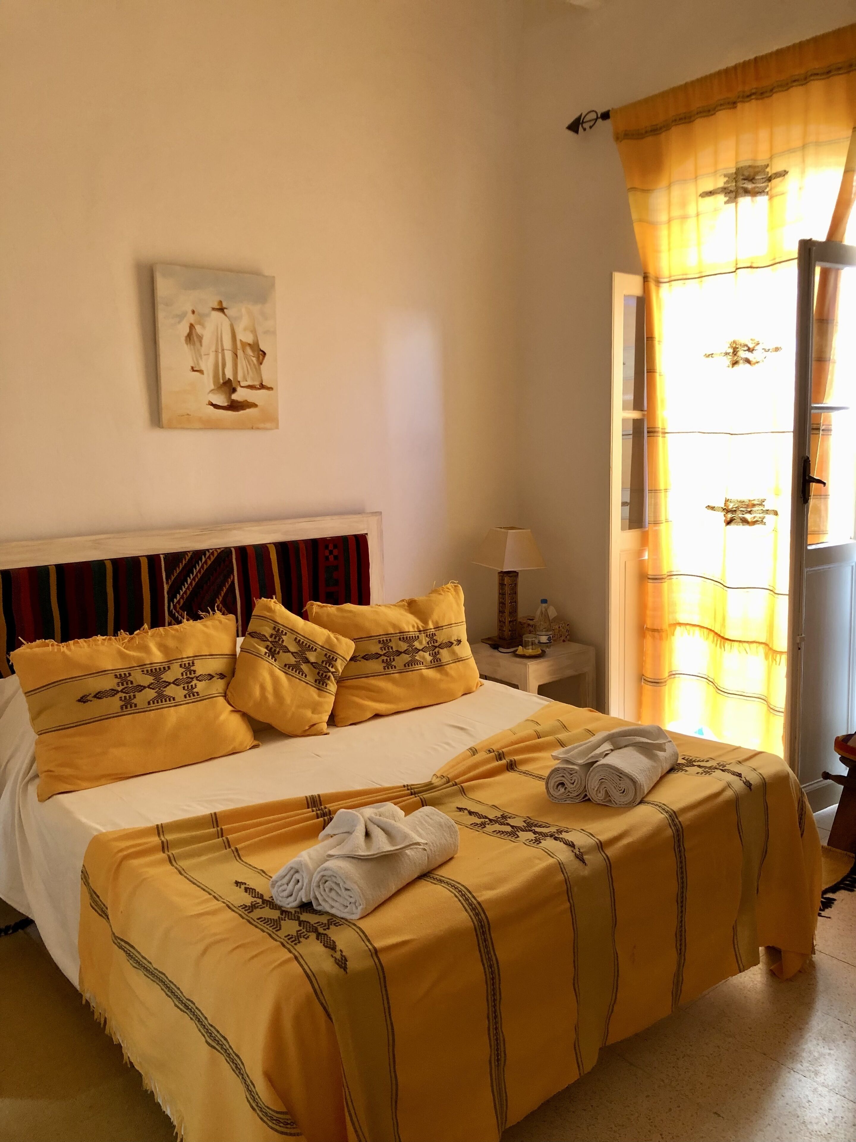 Una cama bien hecha con ropa de cama y almohadas amarillas, un cuadro en la pared y una ventana con cortinas.