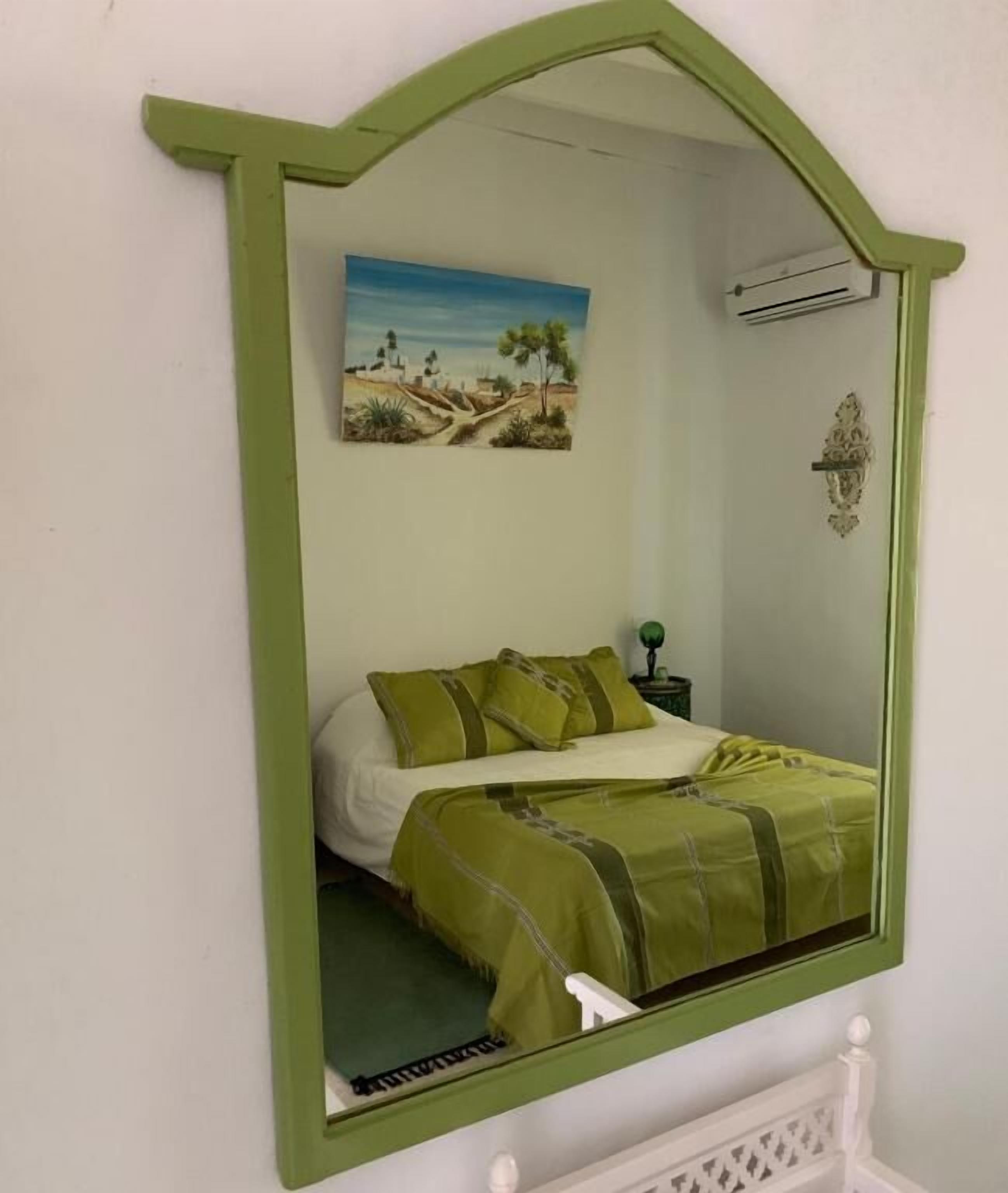 A bedroom with a bed, green bedding, a painting of a landscape, an air conditioning unit, and a decorative wall piece.