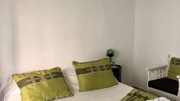 Een net opgemaakt bed met groene beddengoed en kussens, een schilderij van een landschap aan de muur en een stoel met een groene kussen.