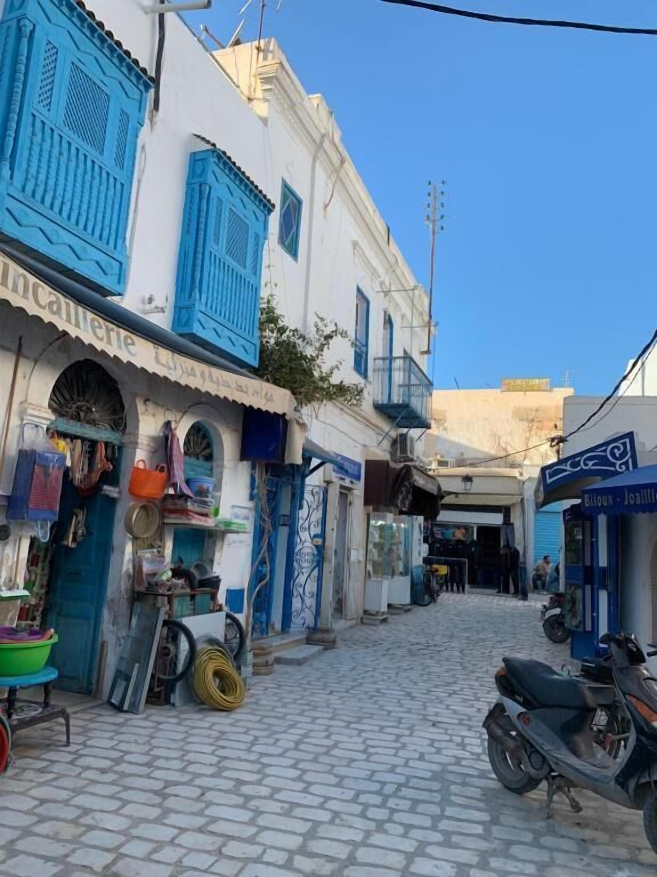 Une rue pavée bordée de boutiques des deux côtés, dont une devanture bien visible avec une enseigne indiquant « Boulouk - Joalil ».