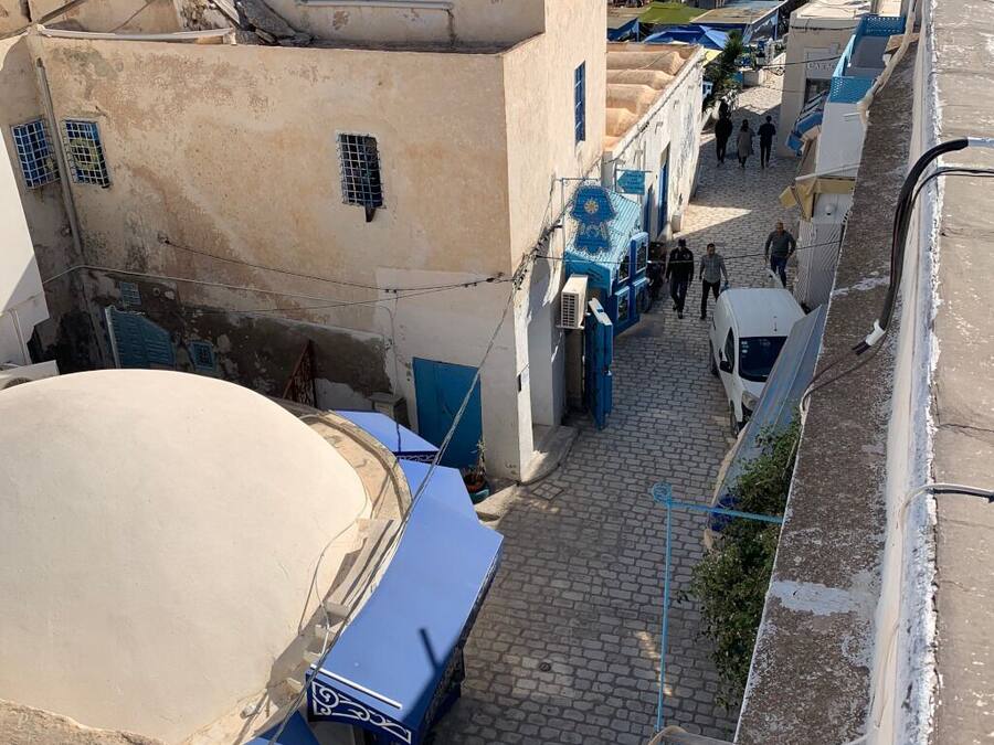 Une rue étroite, bordée de bâtiments des deux côtés, dont certains sont dotés d'auvents bleus et quelques-uns arborent des enseignes bien visibles.