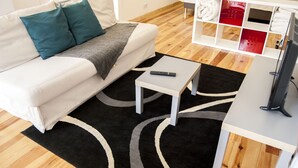 Una sala de estar moderna con un sofá blanco, cojines color esmeralda, una manta gris, un tapete negro y gris, una pequeña mesa de centro blanca y un mueble de TV con televisor de pantalla plana.