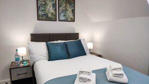 Una cama bien tendida con ropa de cama blanca y aguamarina, dos almohadas y dos toallas dobladas sobre la cama. Hay dos mesitas de noche, una con una lámpara y una botella de agua.