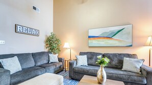 Ein Wohnzimmer mit zwei grauen Sofas, einem beigen Hocker, einem Holztisch, einer Stehlampe, einer Topfpflanze und einem an der Wand hängenden abstrakten Gemälde.