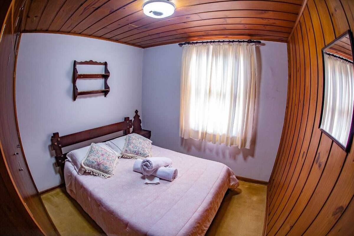 A bedroom with a wooden ceiling, a bed with a pink bedspread, a bedside table with a lamp, and a window with curtains.
