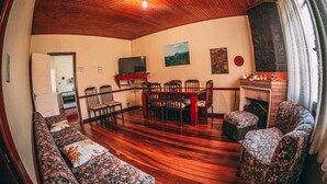A cozy living room with wooden flooring, a sofa set, a dining table with chairs, a TV, and a fireplace. - Colônia de Férias SAA (Campos do Jordão)