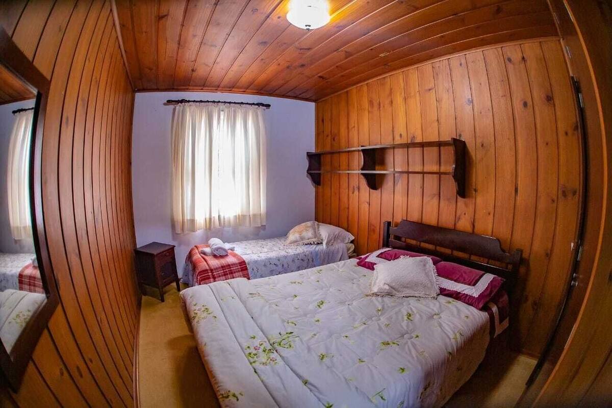 A wooden-paneled room with two beds, a window with curtains, and a shelf on the wall.