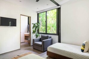 A modern bedroom with a bed, a grey sofa, a TV mounted on the wall, and a large window offering a view of greenery.