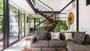 Una sala de estar moderna con un sofá gris, cojines estampados, una mesa de centro de madera y una escalera que lleva a otro nivel.