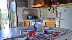 Une cuisine avec un mur en pierre, un plafond en bois, un réfrigérateur blanc et une table sur laquelle on trouve des bols colorés et une bouteille d’eau.
