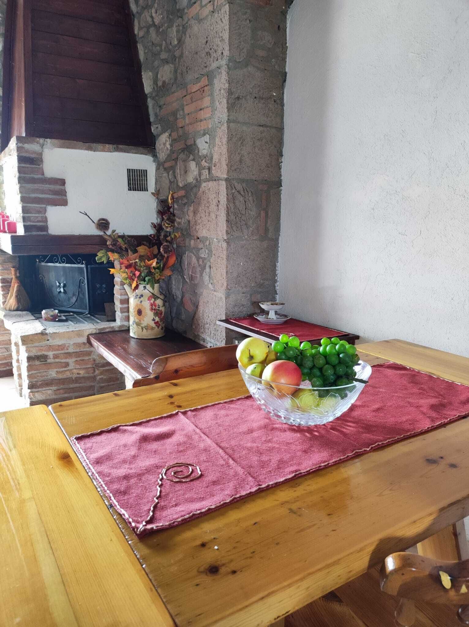 Una mesa de madera con un bol de frutas de vidrio, un paño rojo y una pared de ladrillo con un pilar de piedra.