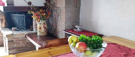 Una mesa de madera con un bol de frutas de vidrio, un paño rojo y una pared de ladrillo con un pilar de piedra.