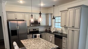 Una cocina moderna con armarios blancos, un refrigerador de acero inoxidable, una isla de granito con taburetes de bar, iluminación colgante y un horno empotrado.