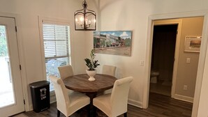 Un comedor con una mesa redonda, cuatro sillas, un florero con flores, un cuadro en la pared y un pasillo que conduce a otra habitación.