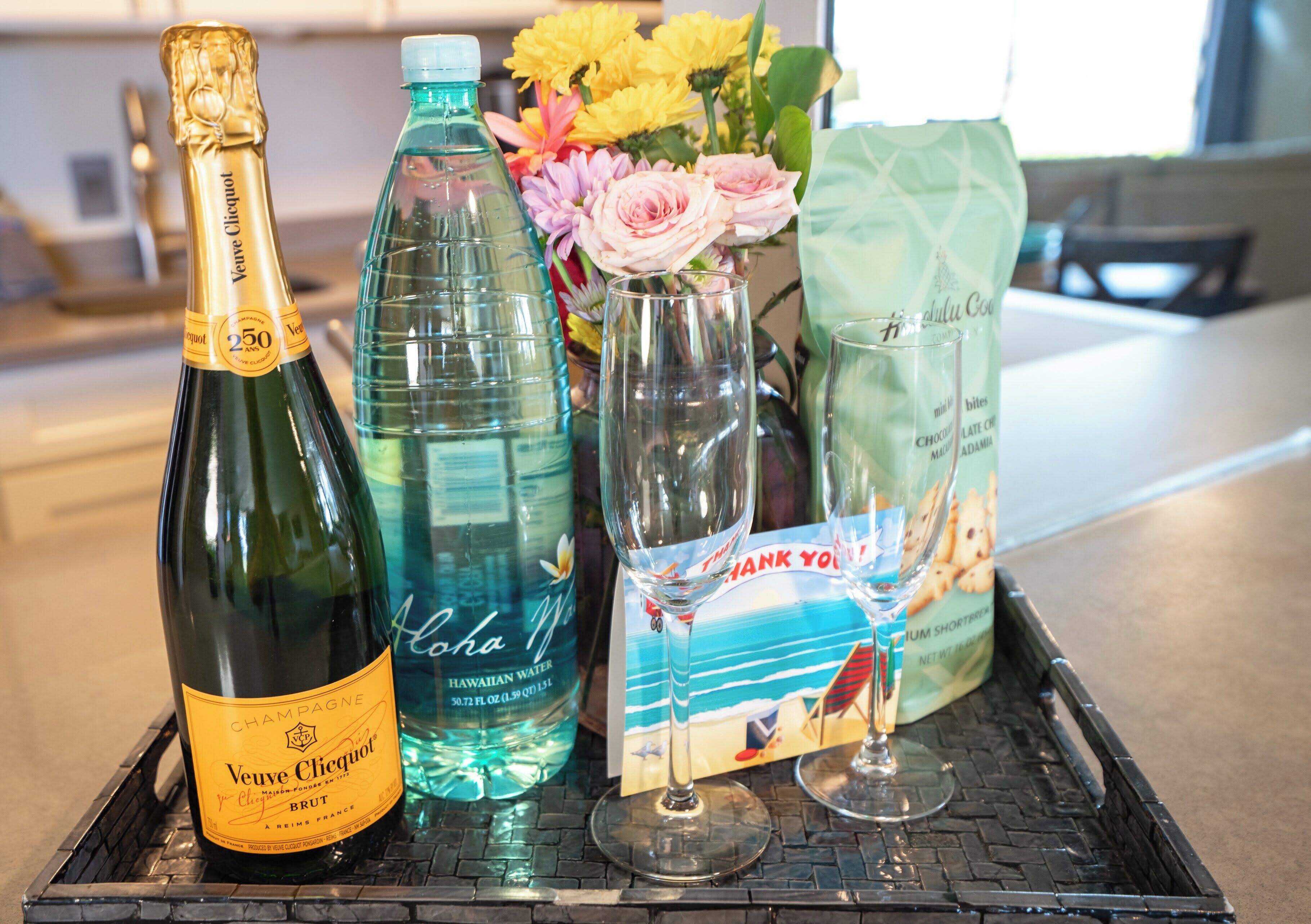 Ein Tablett mit einer Flasche Veuve Clicquot Champagner, einer Flasche hawaiianischem Wasser, zwei Gläsern, einer Dankeskarte und einer Vase mit Blumen.
