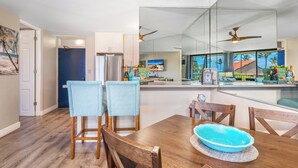 A modern kitchen with a large island, wooden chairs, and a dining table with a blue bowl. - Sunny and Soothing Oceanfront Vistas (Kihei)