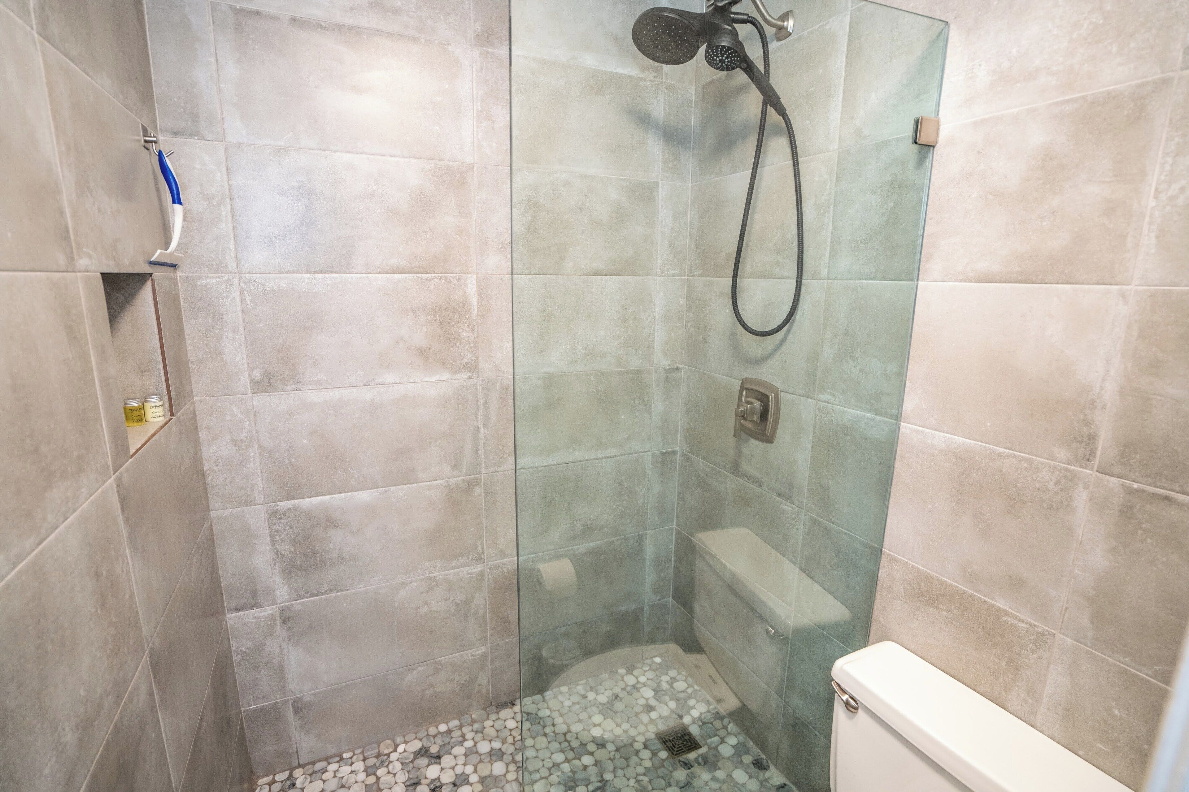 Un vestuario con ducha envuelta en vidrio, cabezal de ducha manual, inodoro y piso de azulejos.