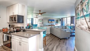 A modern kitchen with a microwave, oven, and a dining area with a table and chairs. There is a living area with a sofa and a TV mounted on the wall. - Sunny and Soothing Oceanfront Vistas (Kihei)