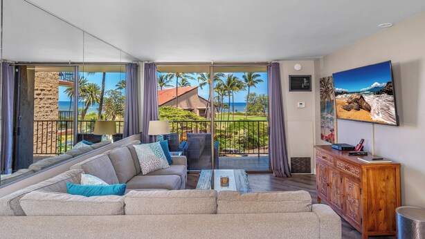 A spacious living room with a large sofa, a coffee table, and a wooden cabinet. A large window offers a view of palm trees and a building. - Sunny and Soothing Oceanfront Vistas (Kihei)