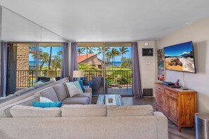 A spacious living room with a large sofa, a coffee table, and a wooden cabinet. A large window offers a view of palm trees and a building. - Sunny and Soothing Oceanfront Vistas (Kihei)