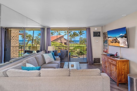 A spacious living room with a large sofa, a coffee table, and a wooden cabinet. A large window offers a view of palm trees and a building.
