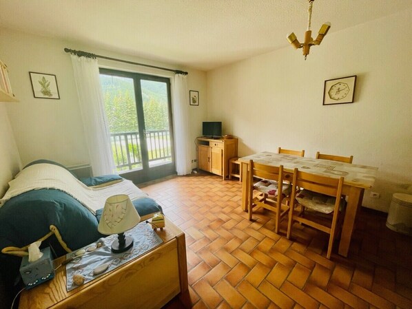 A bedroom with a bed, a bedside table, a lamp, a wooden chair, a small table, a window with curtains, a wooden cabinet, and a wall clock. - 30 m² next to Les Grands Bains (Le Monêtier-les-Bains)