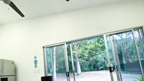 Une chambre moderne avec une grande table en bois, des ventilateurs de plafond et une vue sur une forêt, visible à travers de grandes portes vitrées.