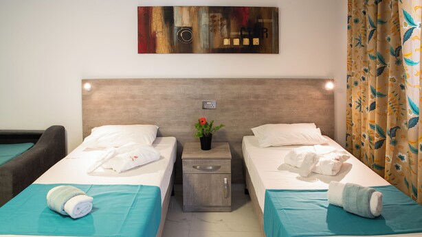 A hotel room with two single beds, a nightstand with a vase of flowers, and a painting on the wall. - Comfort Studio Apartment Combined Living, Bedroom and Kitchen as seen in photo (San Pawl il-Bahar)