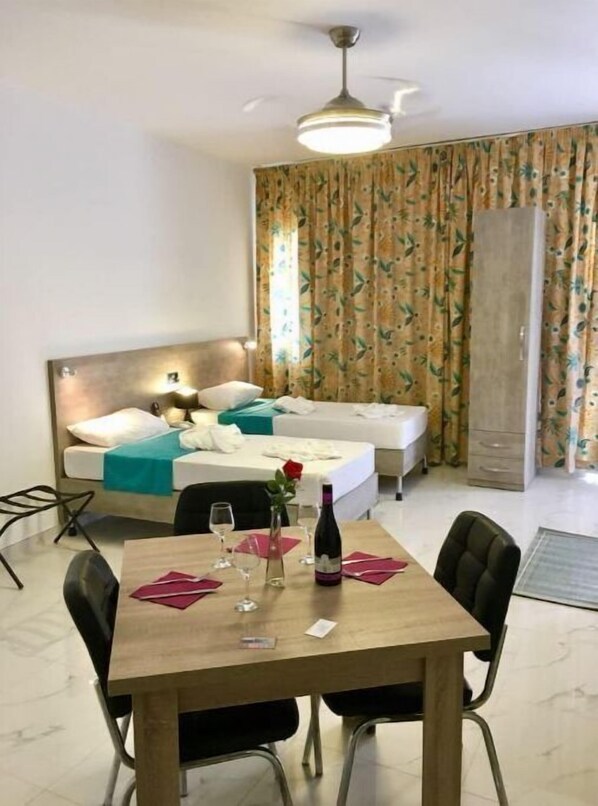 Una habitación de hotel con una cama, una mesa de comedor con sillas, un jarrón con una rosa y una botella de vino.