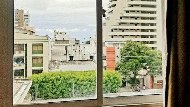 Blick durch ein Fenster auf eine Stadtansicht mit mehreren Gebäuden, darunter ein hohes, weißes Gebäude mit Balkonen und ein niedrigeres Gebäude mit einem begrünten Dach.