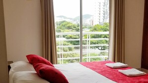 Ein Hotelzimmer mit Bett, einem fensterbeprägten Ausblick auf Gebäude und Grünflächen, sowie einer Fernbedienung auf einem Tisch.
