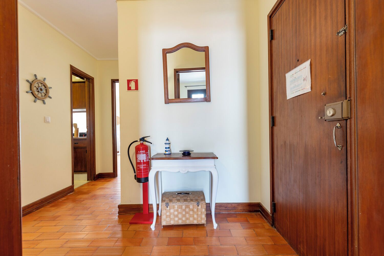 Un pasillo con una puerta de madera, una consola blanca con un espejo encima y un extintor, además de un pequeño aparador.
