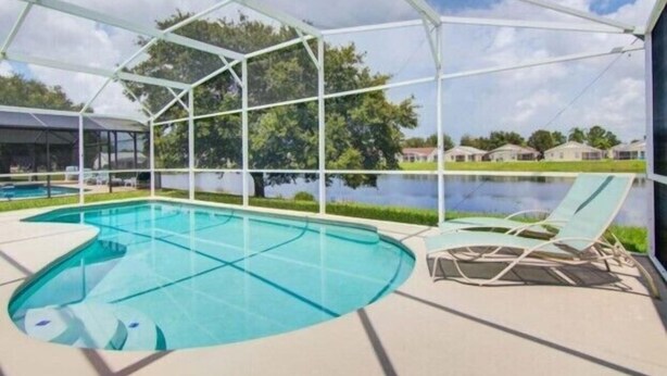 A swimming pool with a covered area, surrounded by a fence, and a view of a lake and houses in the background. - Lake View Villa. Peaceful, 3 bed family home away from home. (Clermont)