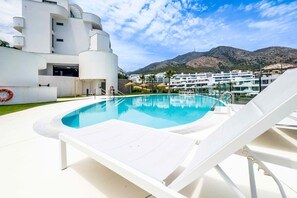 Et moderne poolområde med en klar, blå pool, hvide liggestole og udsigt til et bjerg og hvide bygninger i baggrunden.