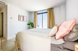 Et hotelværelse med en stor seng, beige gardiner, en potteplante og et vindue med udsigt over byen.