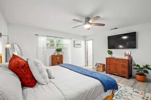 A bedroom with a bed, a ceiling fan, a TV, a dresser, and a window with curtains.
