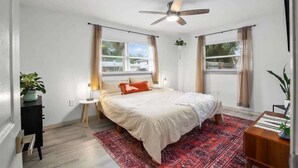 Un dormitorio con cama, ventilador de techo, dos ventanas con cortinas, una mesita de noche pequeña y un aparador.